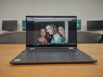 A laptop sits on a particle board table. The background includes desktop computers with the FCC screensaver. The laptop screen shows an AI generated picture of three uncanny young women.